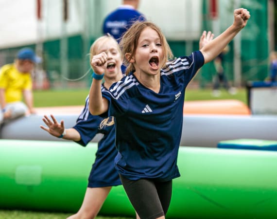 Two young girls in navy sports uniforms joyfully run and cheer outdoors on a grassy field, with inflatable obstacles and other children in the background.