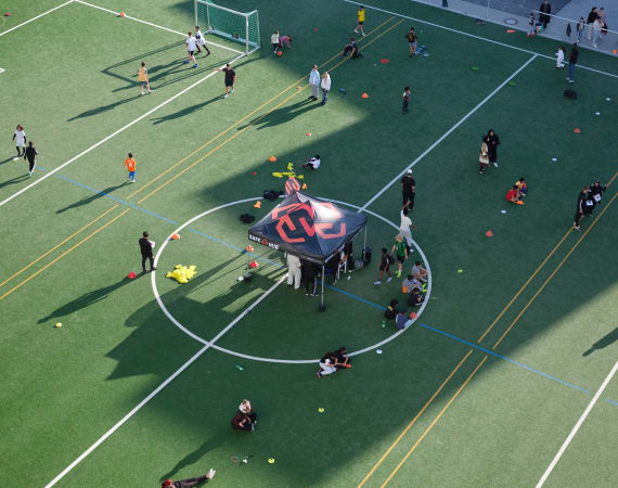 An aerial view of an outdoor sports field where children and adults are spread out participating in various football activities. A small event tent is set up at the center circle, with groups gathered around it. Cones, balls, and training equipment are scattered across the turf, and long shadows stretch across the field on a sunny day.