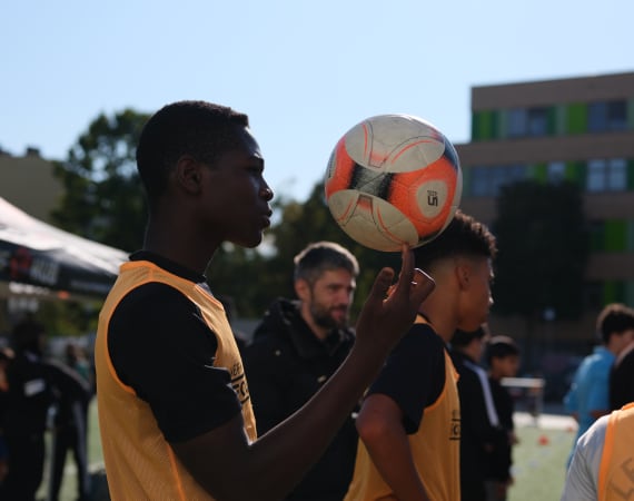 A young person wearing a yellow training vest balances a football on one finger on an outdoor sports field, with other participants and coaches standing nearby. The scene is brightly lit by the sun, and a building with colorful panels is visible in the background.