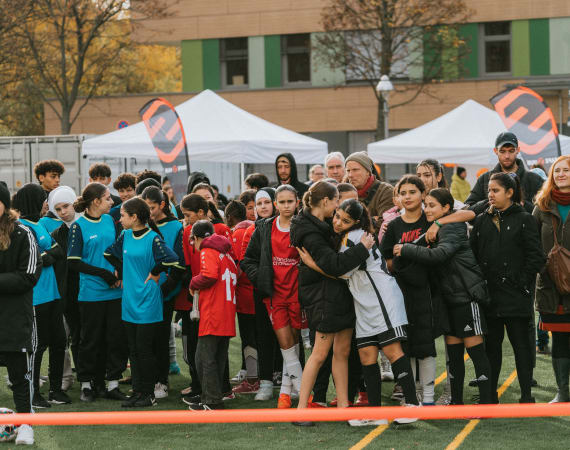 A large group of children and adults gather on an outdoor sports field during an event, with participants wearing sports kits and warm jackets. Some children stand in teams while others hug or talk in small groups. White event tents, flags, and a building with colorful panels are visible in the background, along with autumn trees.