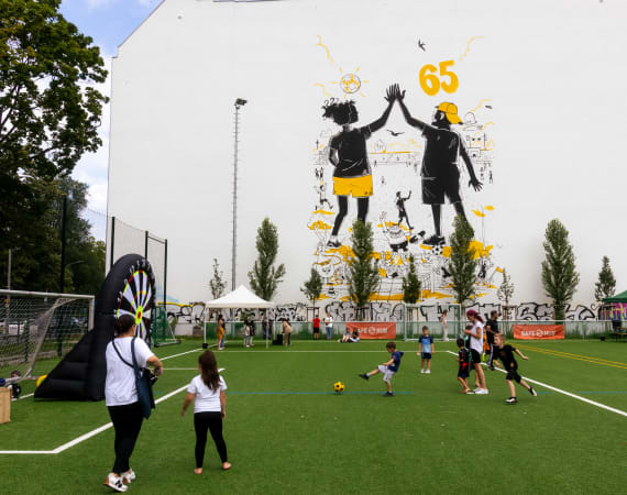 A community sports field with children playing football in the foreground, while adults and other children gather around activity stations. In the background, a large mural on a white building shows two illustrated figures high‑fiving, surrounded by yellow accents and the number 65. Trees, tents, and sports equipment are set up around the field on a sunny day.