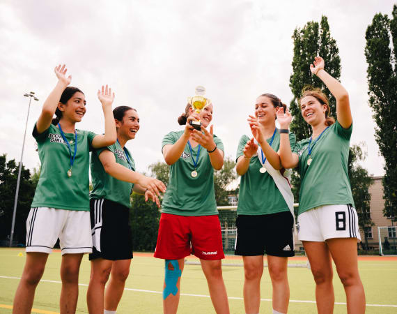 A small football team stands in a circle on an outdoor pitch, cheering as one player lifts a gold trophy. They are wearing green jerseys with medals around their necks, and tall trees and cloudy skies form the backdrop.