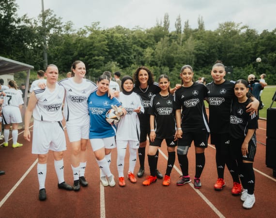 A group of football players in white and black kits stand together on a running track, posing for a team photo. They are wearing matching jerseys, shorts, and socks, and some hold a football. Trees and a green field are visible in the background.