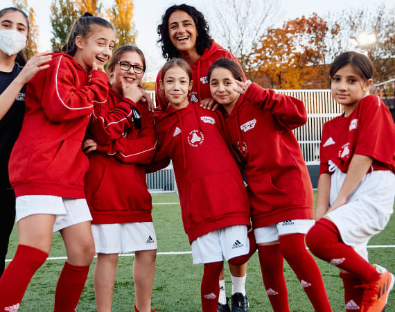 A group of young football players wearing matching red jerseys, shorts, and socks stand together in a playful pose on a sports field. Autumn trees with orange and yellow leaves are visible behind them.