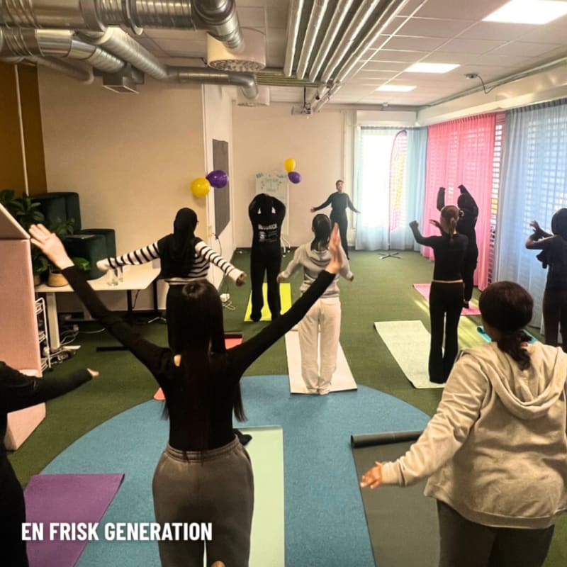 A group of people practice yoga or stretching on mats in a bright room with colorful curtains and balloons. Most have their arms raised.