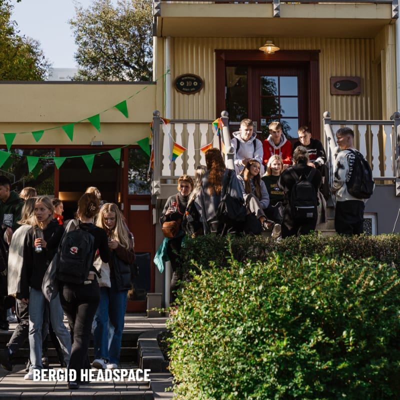 A group of students gathers outside a yellow building decorated with green and orange bunting. Some stand on the steps, others on the ground, talking and smiling in the sunlight.