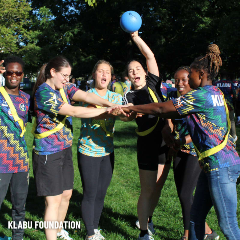A group of people wearing colorful shirts stand in a circle outdoors, placing their hands together, with one person holding a blue ball aloft.