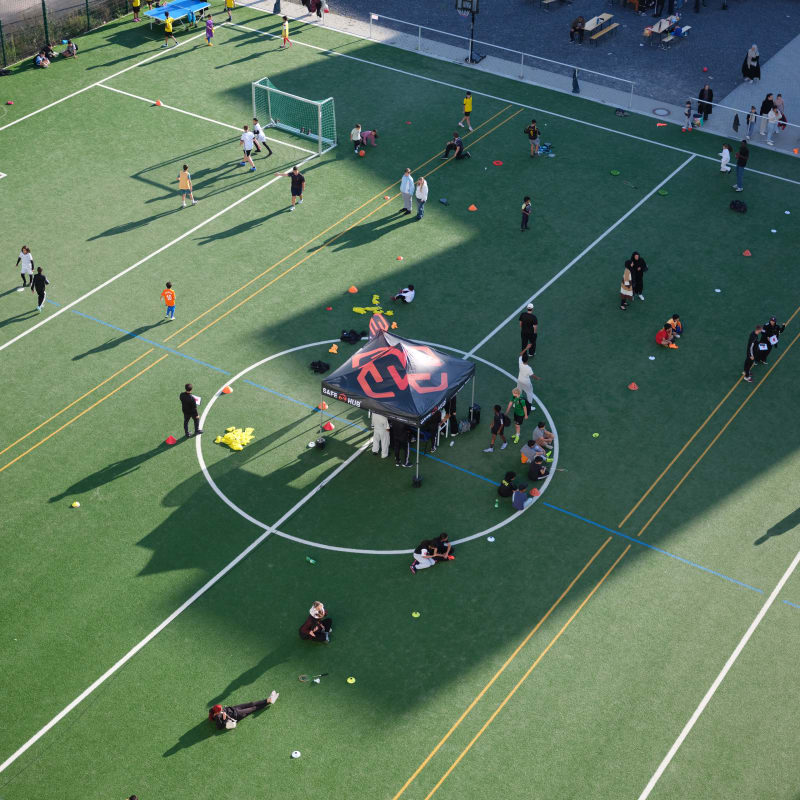 An aerial view of an outdoor sports field where children and adults are spread out participating in various football activities. A small event tent is set up at the center circle, with groups gathered around it. Cones, balls, and training equipment are scattered across the turf, and long shadows stretch across the field on a sunny day.