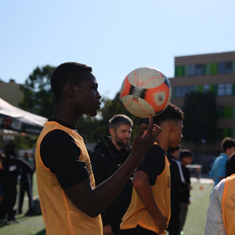 A young person wearing a yellow training vest balances a football on one finger on an outdoor sports field, with other participants and coaches standing nearby. The scene is brightly lit by the sun, and a building with colorful panels is visible in the background.