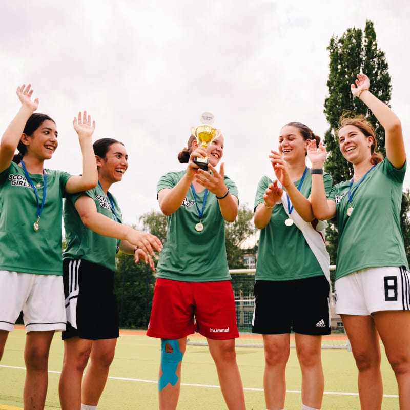 An aerial view of an outdoor sports field where children and adults are spread out participating in various football activities. A small event tent is set up at the center circle, with groups gathered around it. Cones, balls, and training equipment are scattered across the turf, and long shadows stretch across the field on a sunny day.
