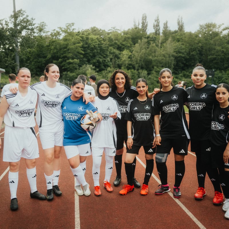 A group of football players in white and black kits stand together on a running track, posing for a team photo. They are wearing matching jerseys, shorts, and socks, and some hold a football. Trees and a green field are visible in the background.