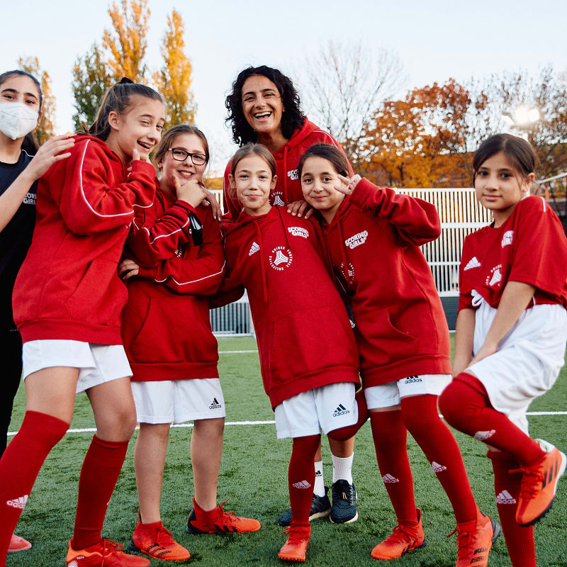 A young person wearing a yellow training vest balances a football on one finger on an outdoor sports field, with other participants and coaches standing nearby. The scene is brightly lit by the sun, and a building with colorful panels is visible in the background.