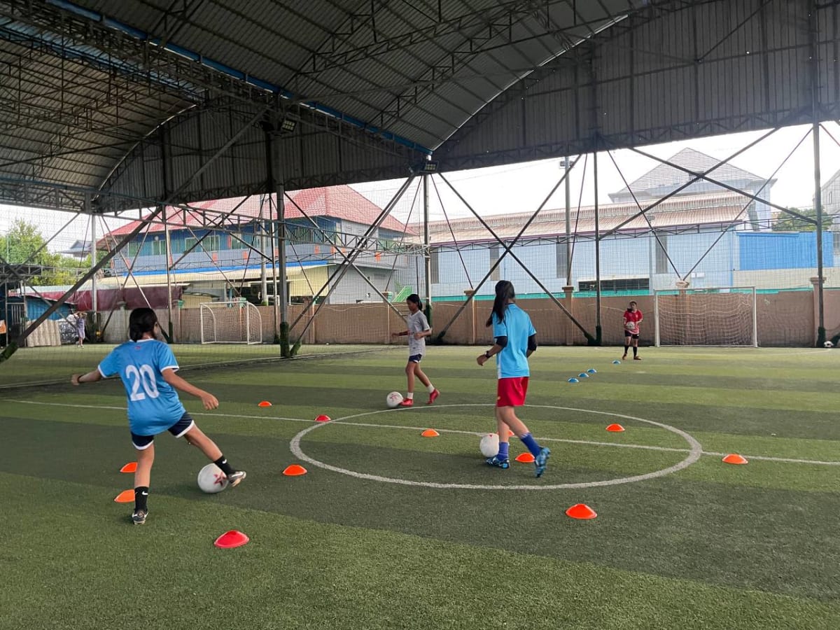 Indoor soccer practice with teenagers wearing blue jerseys. They are dribbling soccer balls around orange cones on a green field, under a metal roof.