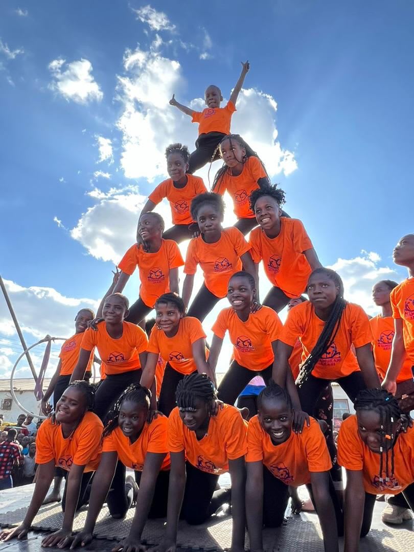 A group of children in bright orange T-shirts form a human pyramid under a sunny blue sky.