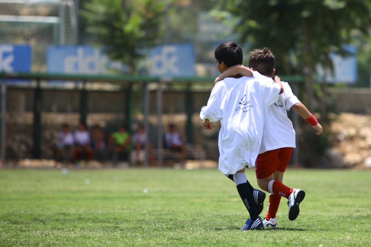 Two young boys wearing sports clothes run together on a grassy field with their arms around each other’s shoulders, suggesting friendship and teamwork, while people sit in the background.