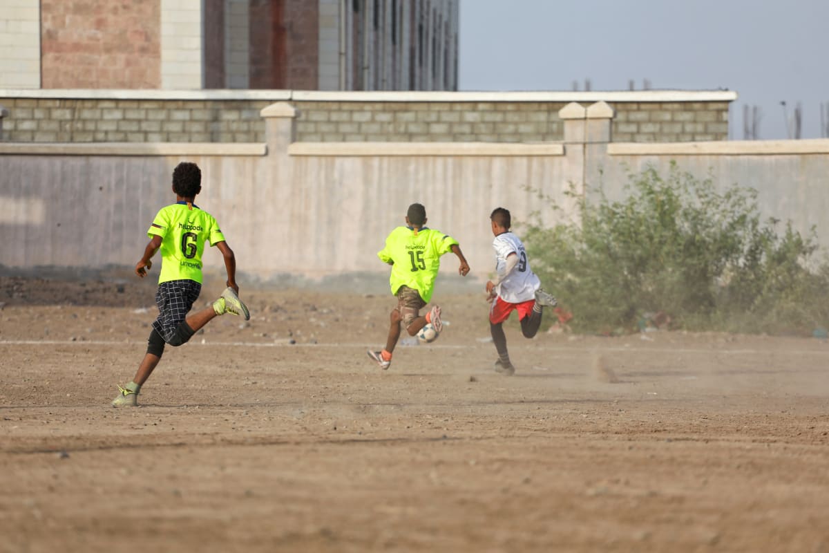 Children playing football on a dusty field in Yemen, with hills and simple buildings in the background. The players wear casual clothes, and the field is unmarked.