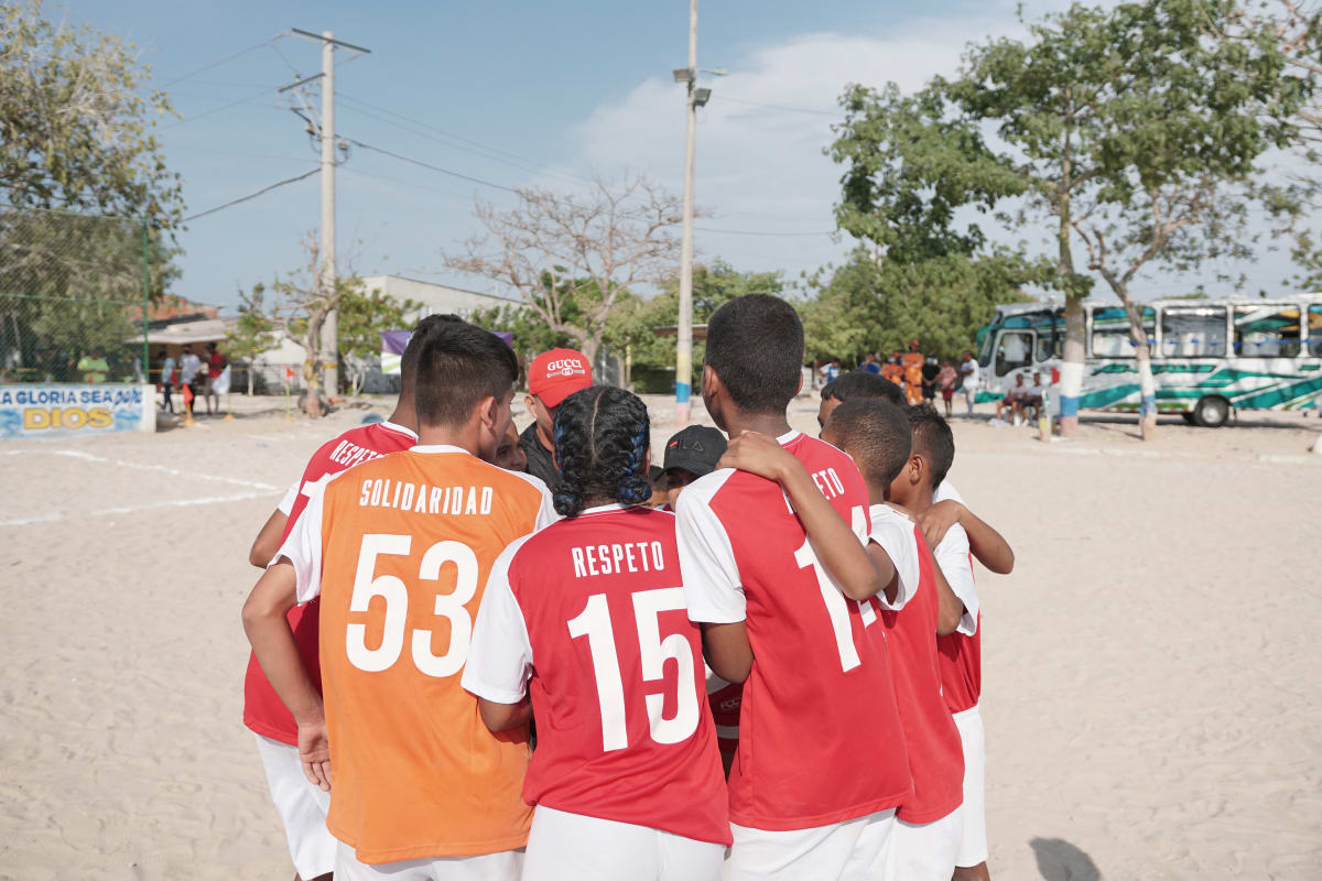 A group of children some in red and some in orange jerseys huddle together on a sandy field. Buses and trees are visible in the background.