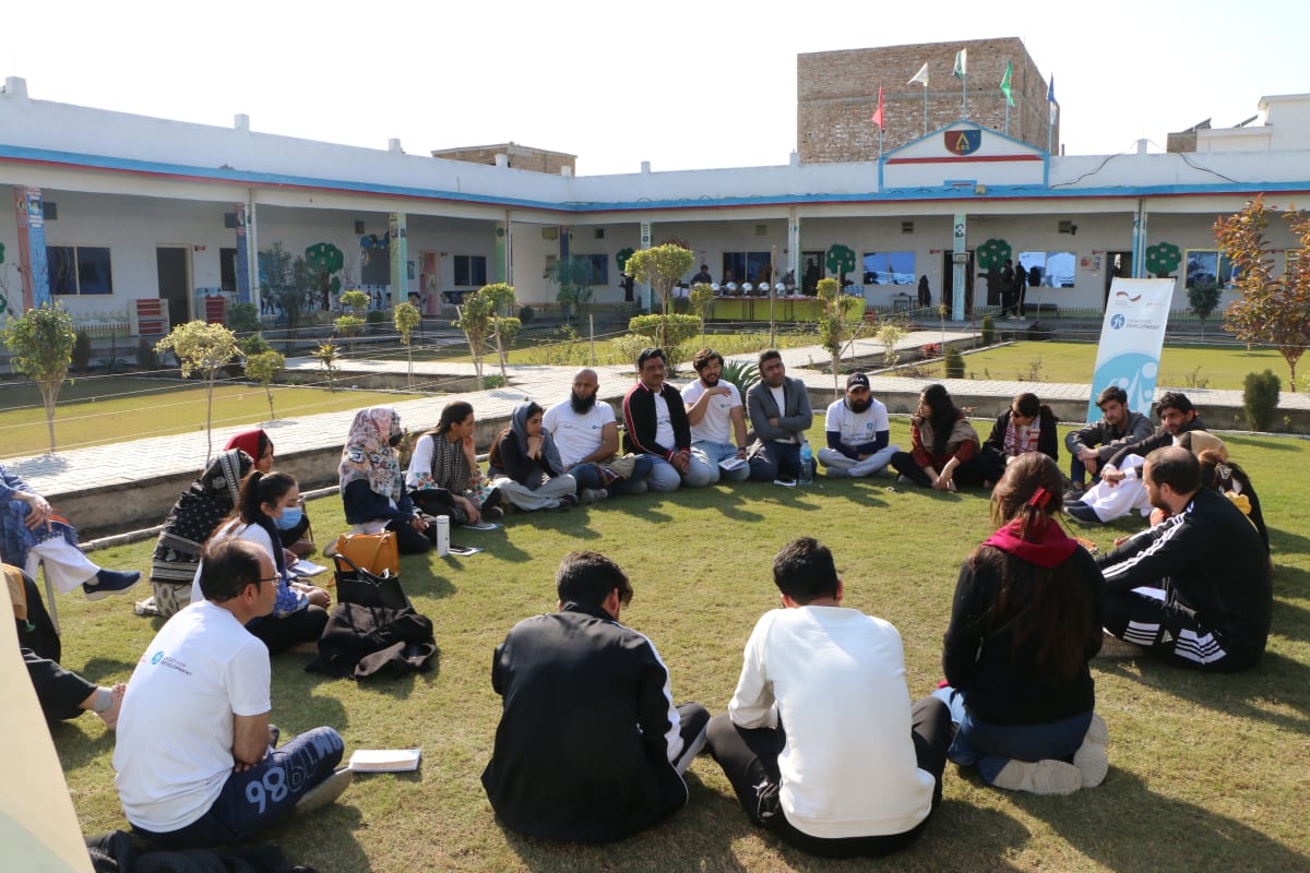 A group of people sits in a large circle on grass in a courtyard, engaged in discussion. The background shows a school building with colorful accents, flags, and small trees. The weather is sunny and clear.