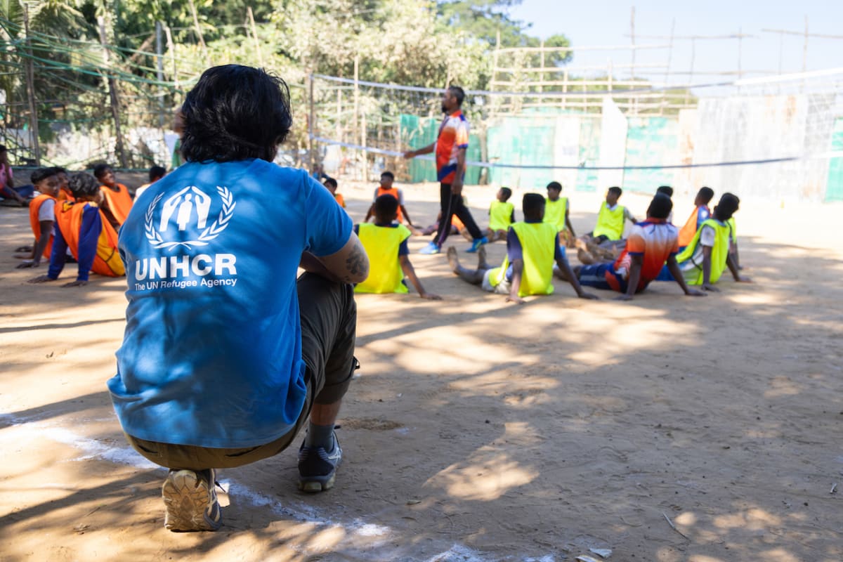 A person wearing a UNHCR shirt crouches near a group of children sitting on the ground in a circle outdoors, as another adult stands in front of them, possibly leading an activity or game.
