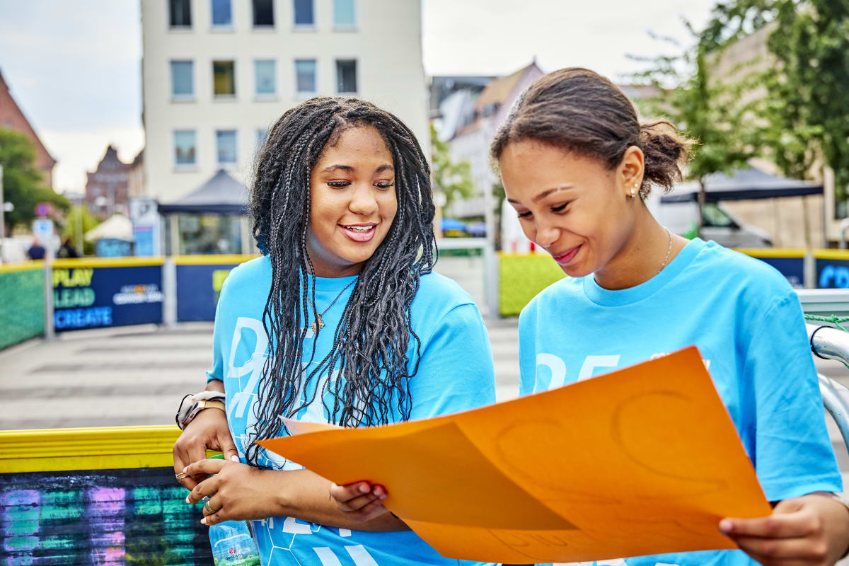 Two women wearing blue shirts smile as they look at an orange poster together. They're outdoors in a lively urban setting.