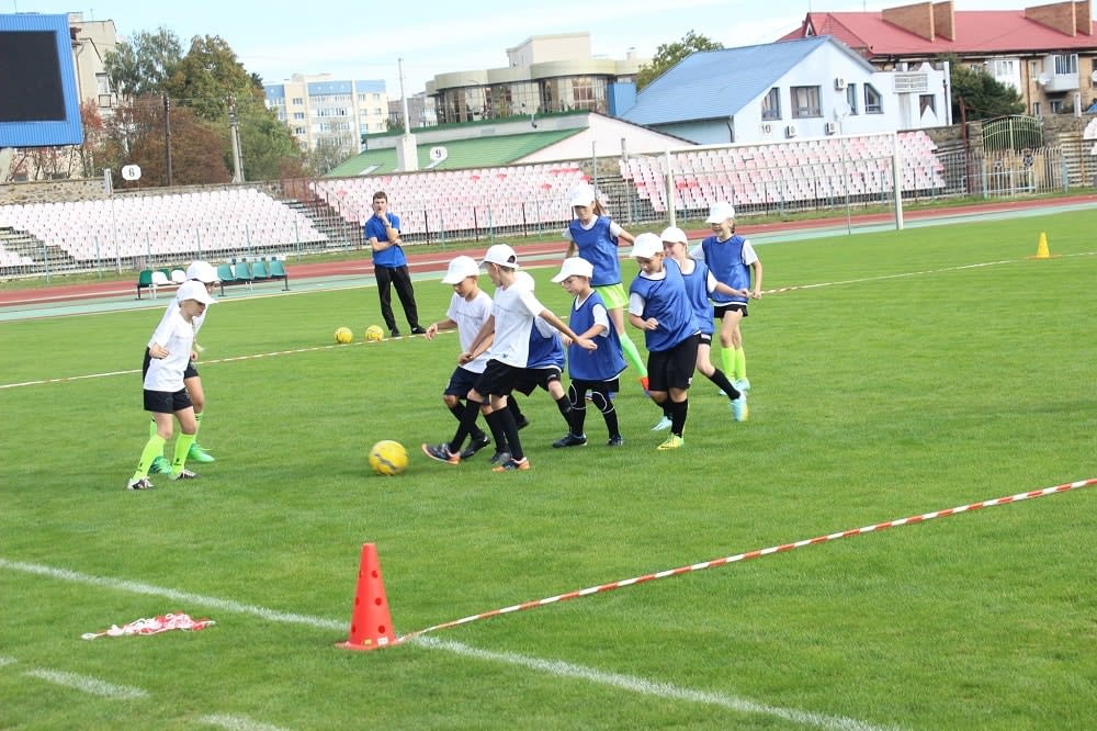 Kids in white and blue jerseys play soccer on a grassy field, surrounded by orange cones. A coach observes from the sidelines.