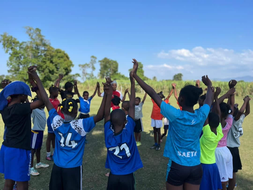 A group of children standing in a circle on a grassy field under a clear blue sky, holding hands with arms raised.
