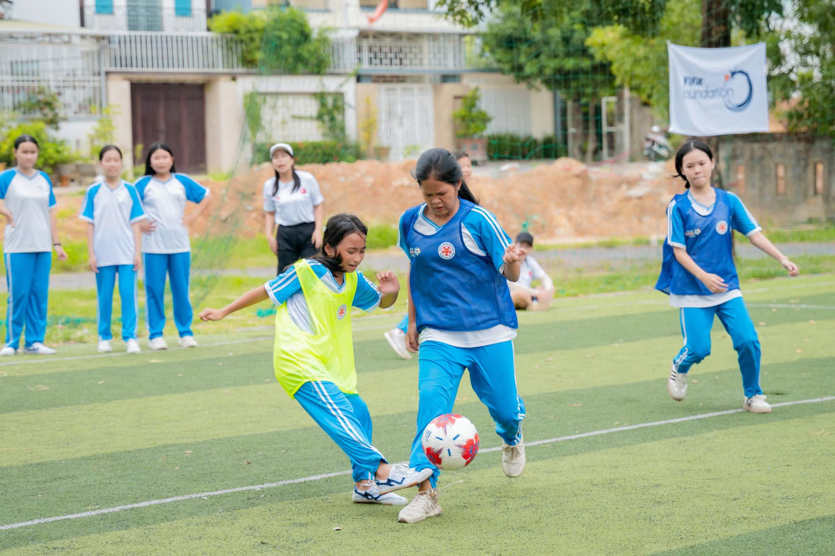 Two girls, one in a yellow vest and another in a blue vest, are playing soccer on a grassy field, focused on the ball. There are other players and observers in the background.