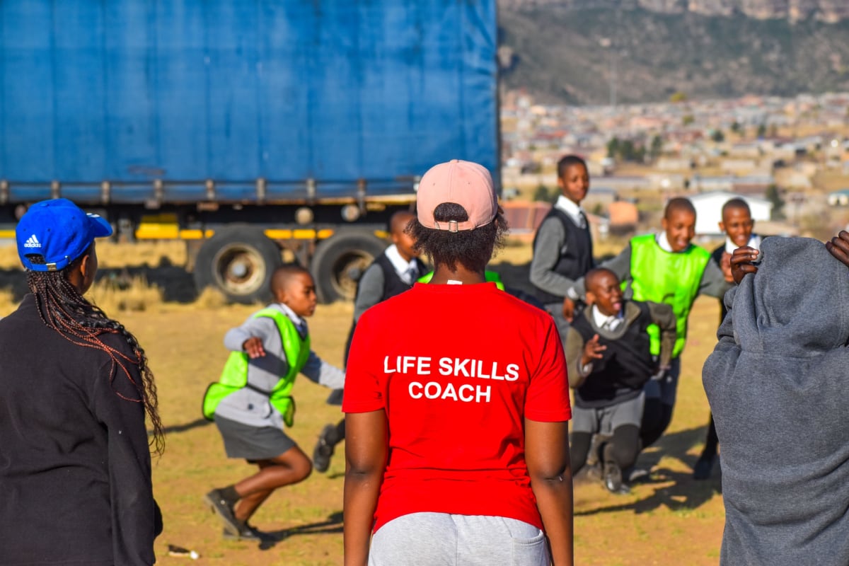 A woman wearing a red shirt labeled LIFE SKILLS COACH watches a group of schoolchildren in uniforms and green vests playing outdoors, with a blue truck and town in the background.