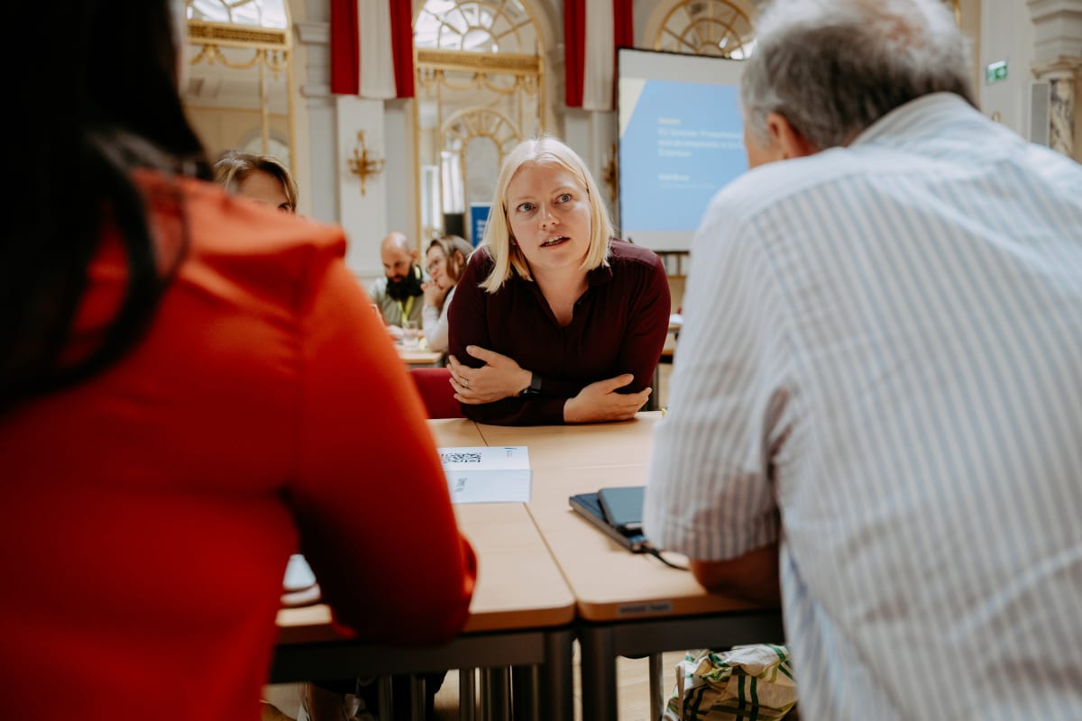 A woman in a maroon shirt leans forward, attentively listening to colleagues in a conference room.