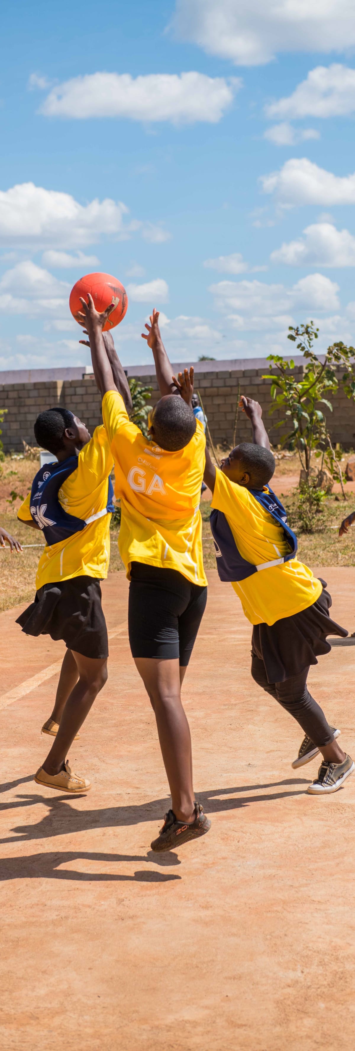 Children wearing yellow and blue sports uniforms jump with arms raised, reaching for an orange ball on an outdoor court under a partly cloudy sky.