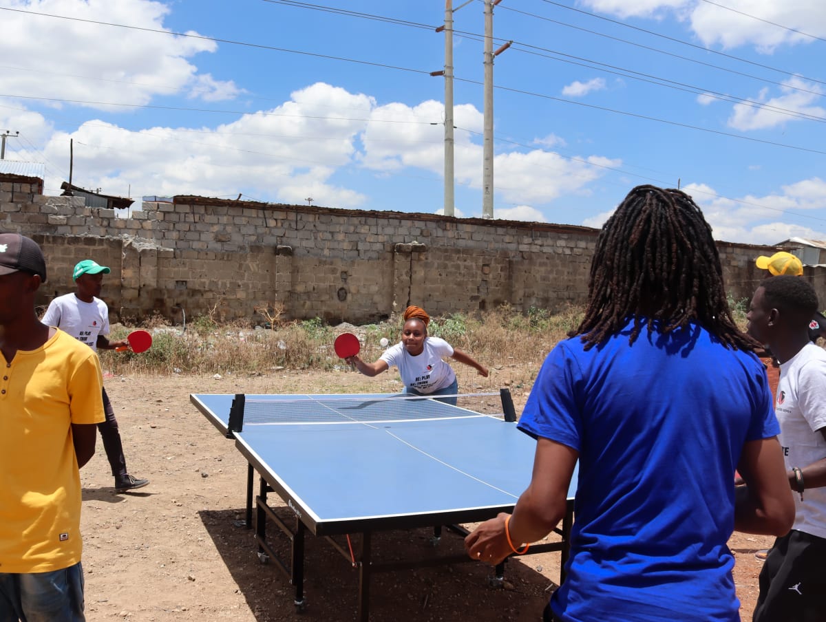 A group of people play table tennis outdoors on a sunny day, surrounded by a stone wall and power lines. One woman is mid-action, swinging her paddle, as others watch or wait their turn.
