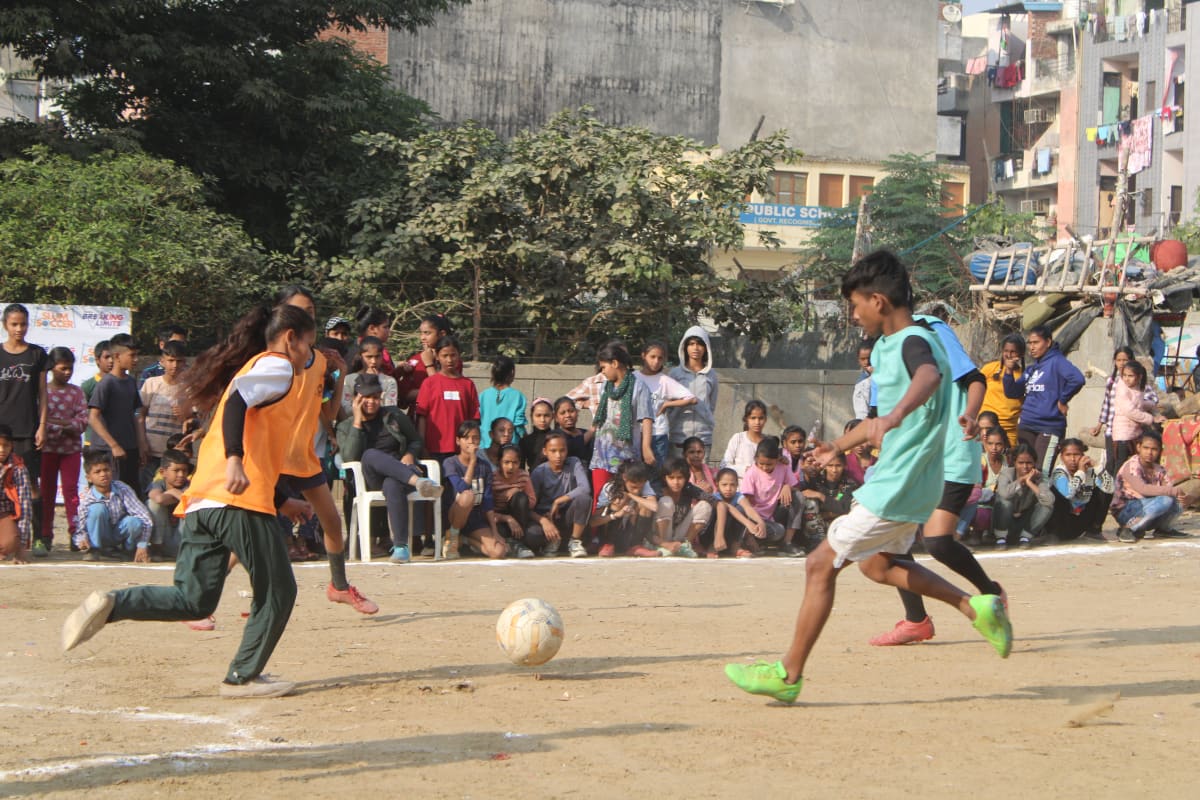 Children play a soccer match on a sandy field while a large crowd of spectators, including kids and adults, watch from the sidelines. Buildings and greenery are visible in the background.
