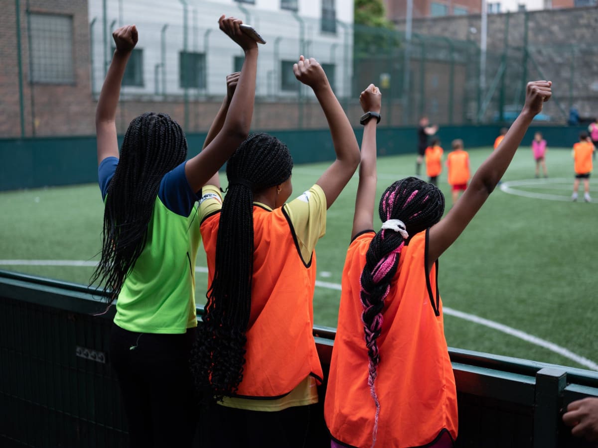 Three girls with braided hair, wearing sports bibs, stand by a fence with arms raised, cheering on a group of children playing soccer on an outdoor field.