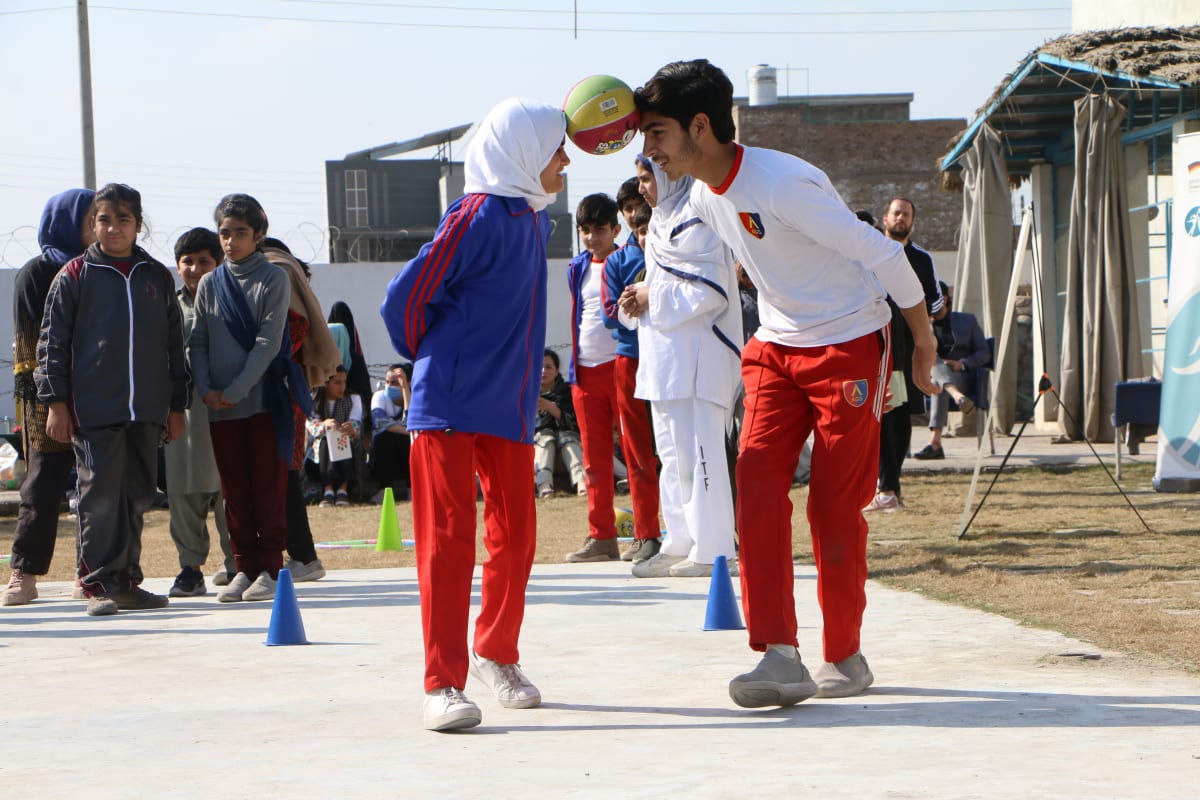 Two children balance a ball between their foreheads during an outdoor activity, surrounded by peers.