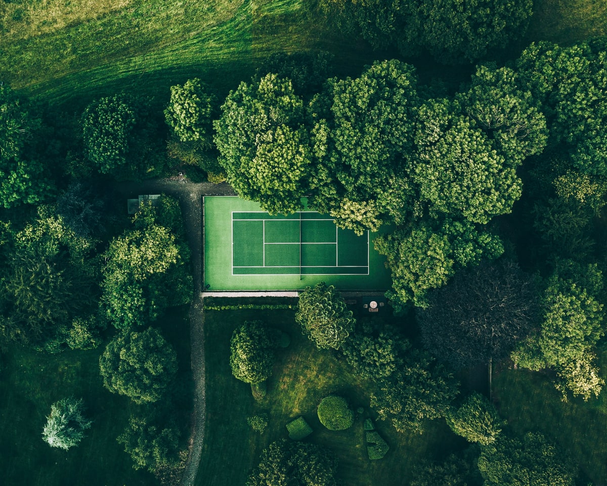 Aerial view of a green tennis court surrounded by dense, leafy trees and lush vegetation, creating a striking contrast between the court and the natural landscape.