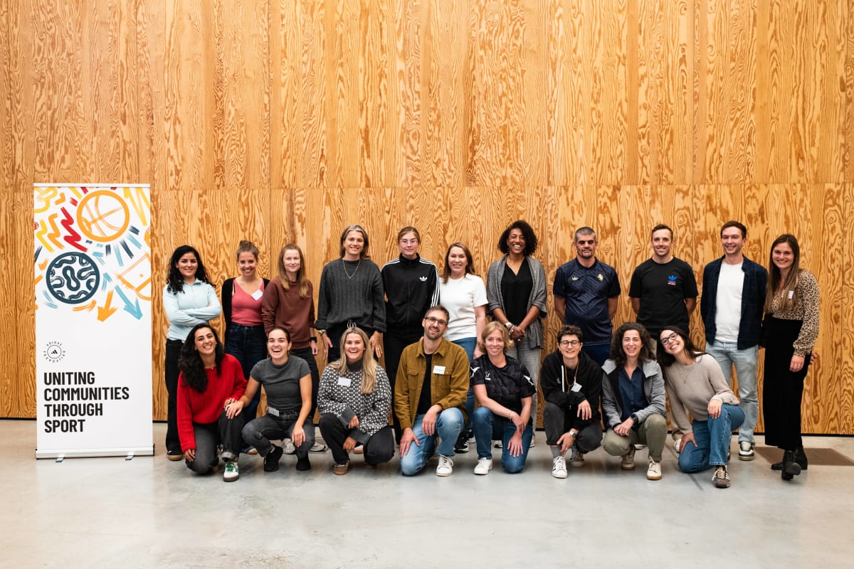 A group of adults posing for a group photo indoors against a wooden wall, with a banner reading “Uniting Communities Through Sport” displayed on the left.