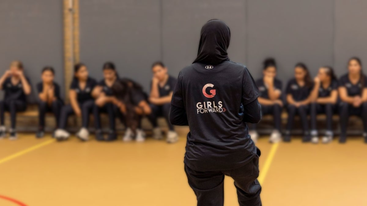 Two women stand in a gym, wearing "Girls Forward" shirts, facing a row of seated individuals.