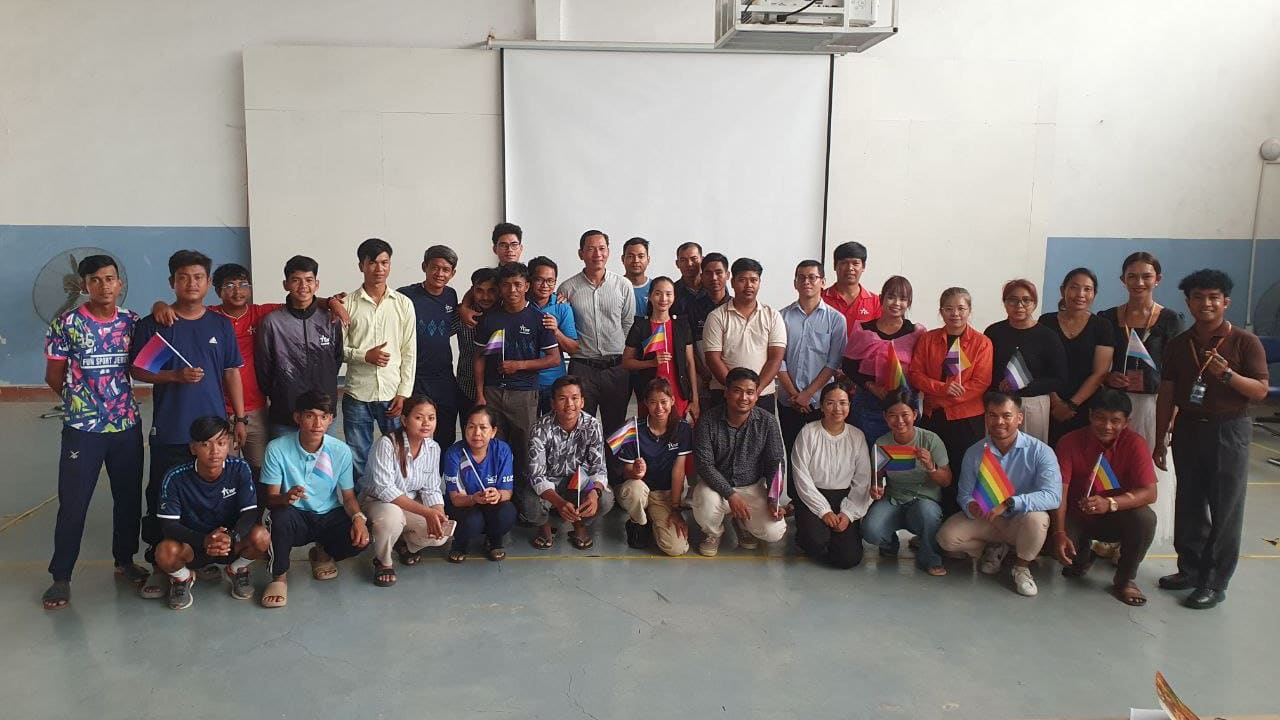 A large group of people poses together indoors in front of a blank projector screen. Some hold rainbow flags and awards. The group appears cheerful and diverse, standing and kneeling in two rows on a blue floor.