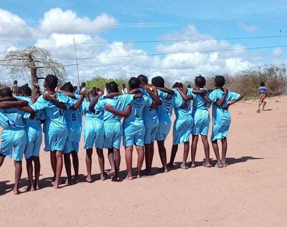 A group of children stand in a line with backs to the camera and arms around each other, wearing light blue soccer uniforms on a sandy field, under a blue sky. Trees and bushes are visible in the background.