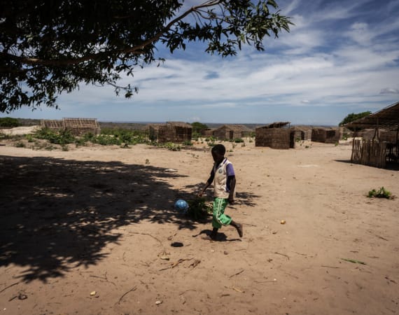 A child plays soccer with a blue ball on sandy ground in a rural village. Straw huts are visible under a blue sky.