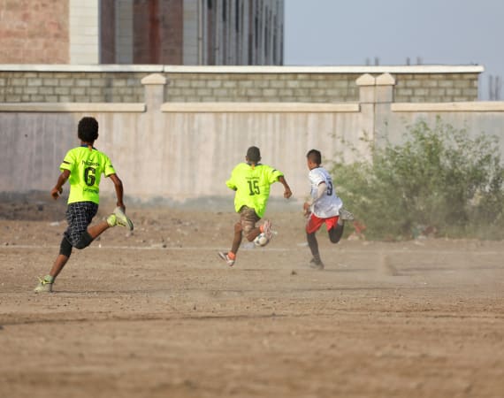 Three young boys play soccer on a dusty field. Two in neon green shirts chase the ball, while one in white shorts is ahead, near a concrete wall.