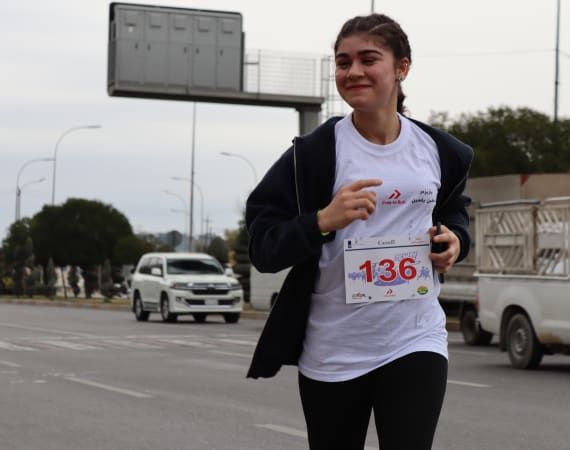 Young woman running a marathon, wearing a white shirt with a race bib number 136, and a black hoodie. She is smiling.