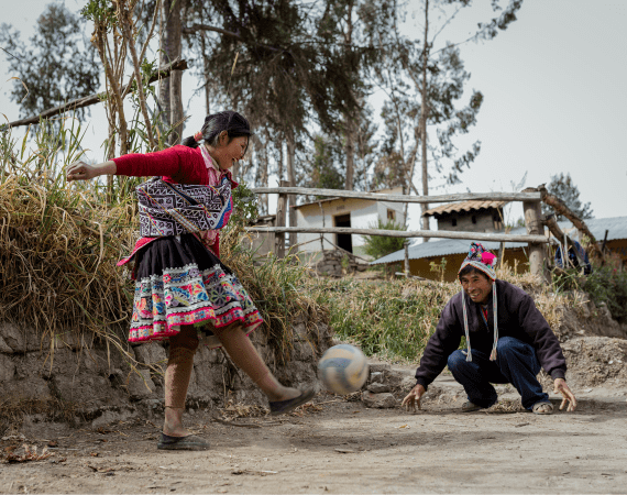 A young girl in traditional clothing kicks a soccer ball toward a smiling man crouching as goalie outdoors in a rural setting with houses and trees in the background.