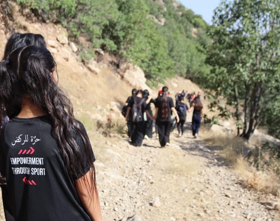 A group of young girls hike along a rocky, tree-lined path in sunlight. A woman in the foreground has "Empowerment Through Sport" text on the back of her black T-shirt.