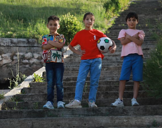 Three children stand confidently on outdoor stone steps. The center child, holding a soccer ball, wears a red shirt. The other two cross their arms and smile. Bright, sunny day with greenery in the background.