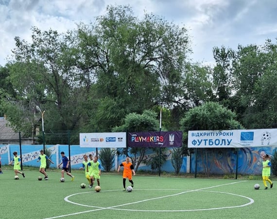 Children in colorful sportswear practice soccer on a lush green field. They're surrounded by trees and banners, under a partly cloudy sky. 