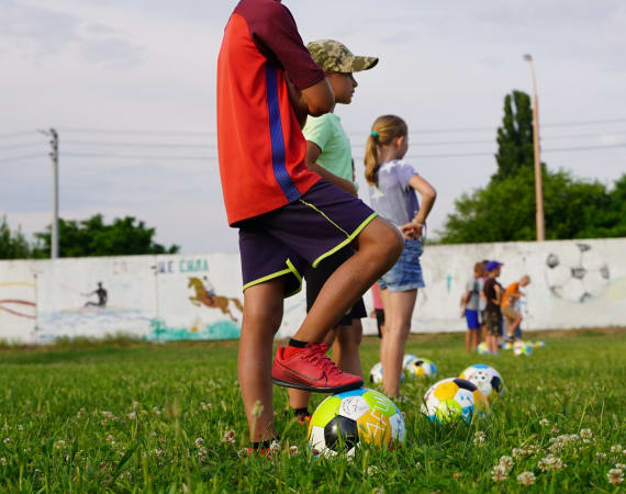 Children stand on grass with colorful soccer balls, one child in a red jersey and cap. They are preparing for a soccer activity.