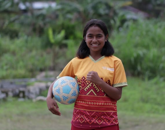 A young girl with dark hair, wearing a yellow and red patterned sports jersey, stands outdoors on grass, smiling and holding a blue and white soccer ball under her arm.