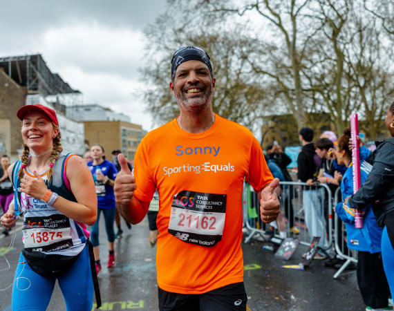 A man running a marathon in a Sporting Equals T-shirt, smiling and showing two thumbs ups.