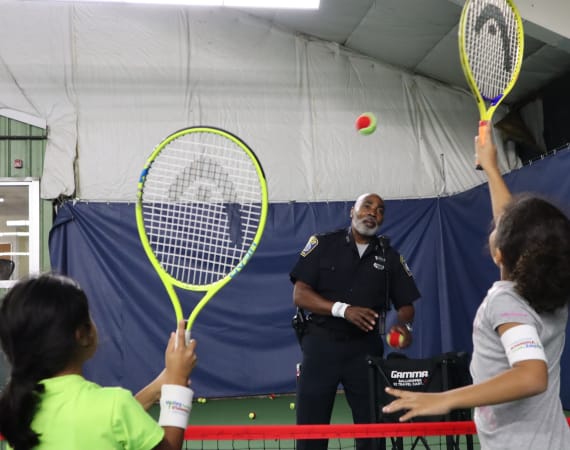 A police officer smiles while tossing tennis balls to two children holding rackets inside an indoor tennis court. The children are reaching up to hit the balls.
